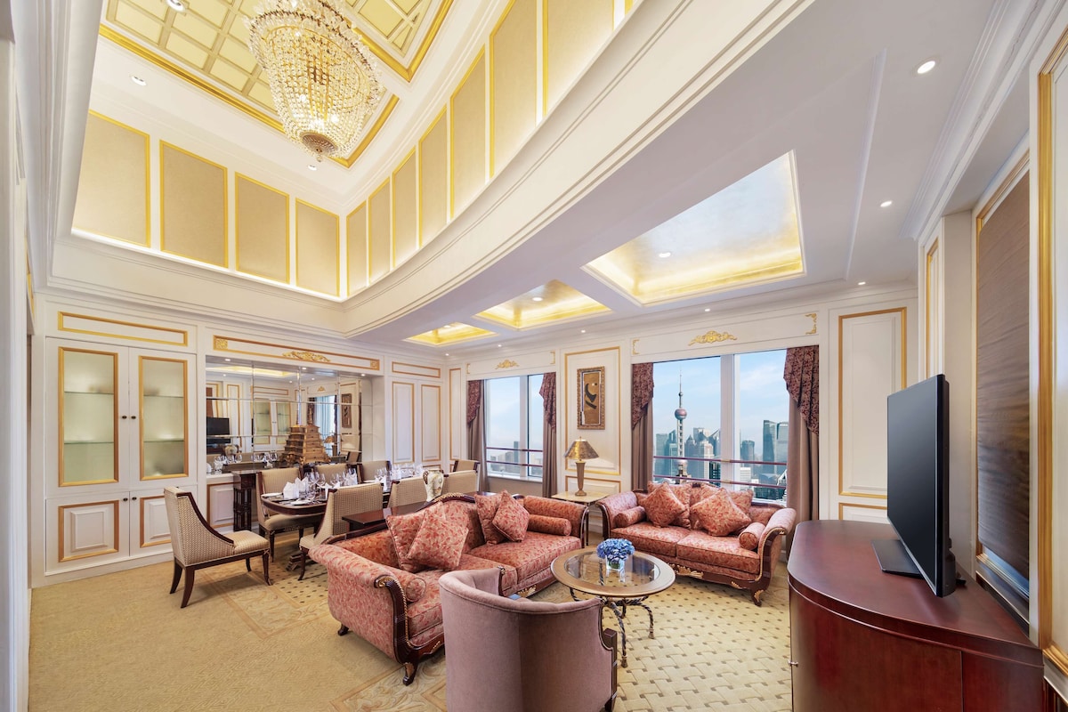A spacious living room with a high ceiling, large windows offering a city view, a dining area with a chandelier, and a modern entertainment unit.