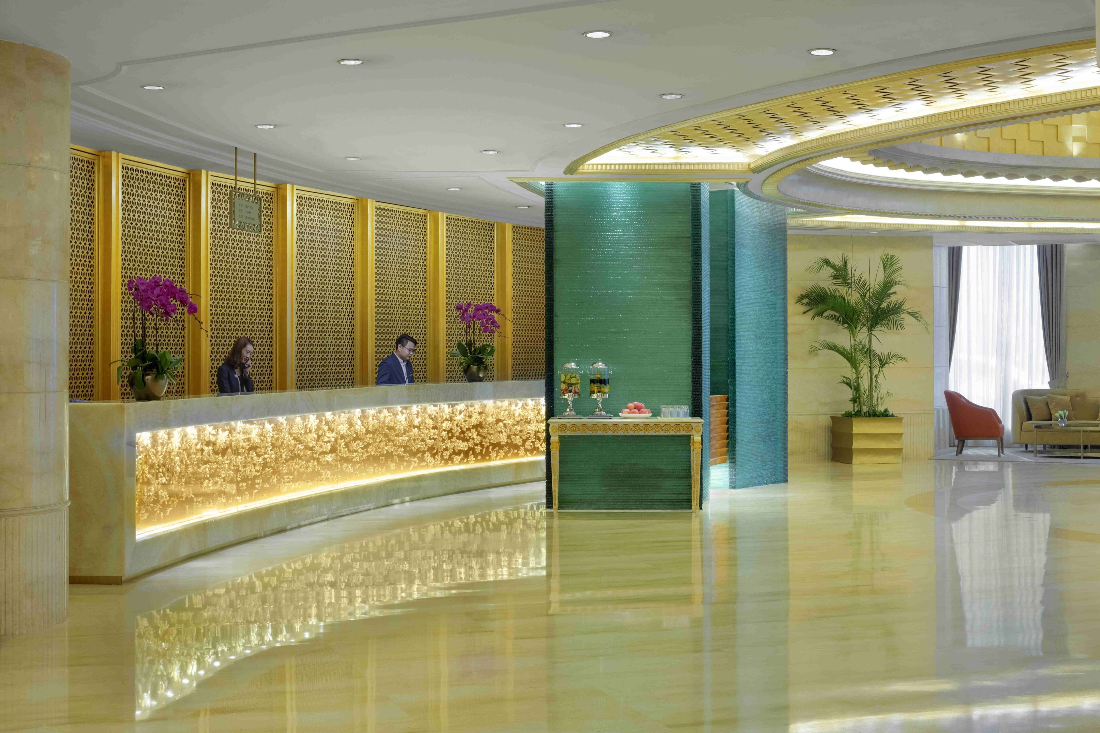 Un moderno área de recepción de hotel con un mostrador curvo, una pared de acento verde y una zona de estar con sofá y sillones.