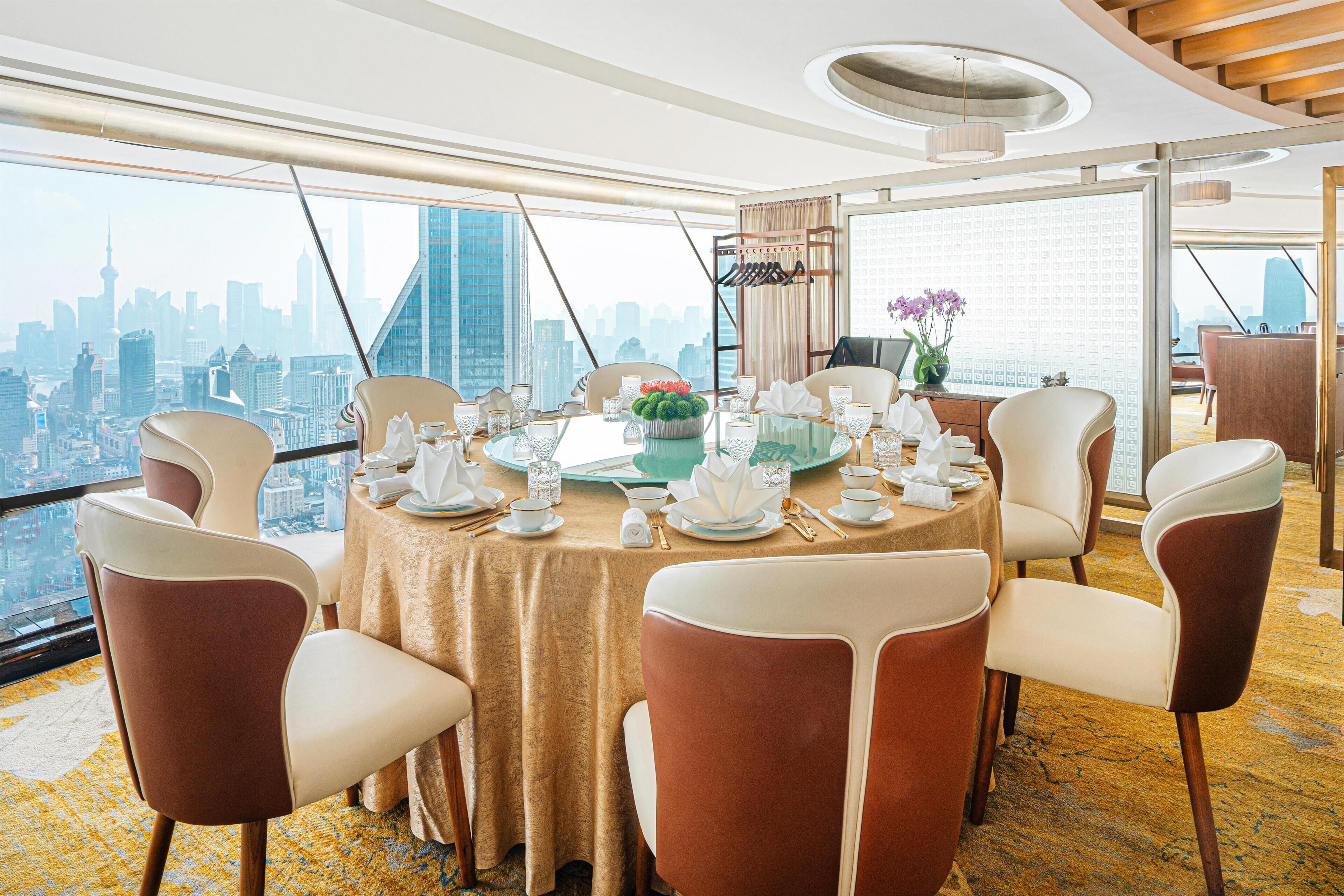 A dining area with a table set for six, large windows offering a city skyline view, and a high ceiling with a circular light fixture.