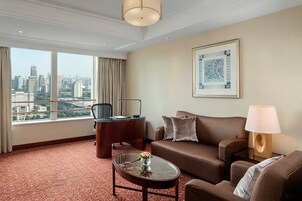 A hotel room with a large window, a sofa, a coffee table, a desk, and a lamp.