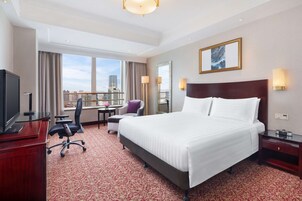 A hotel room with a large bed, a desk with a TV, a chair, a small table with a vase, and a view of the city through large windows.