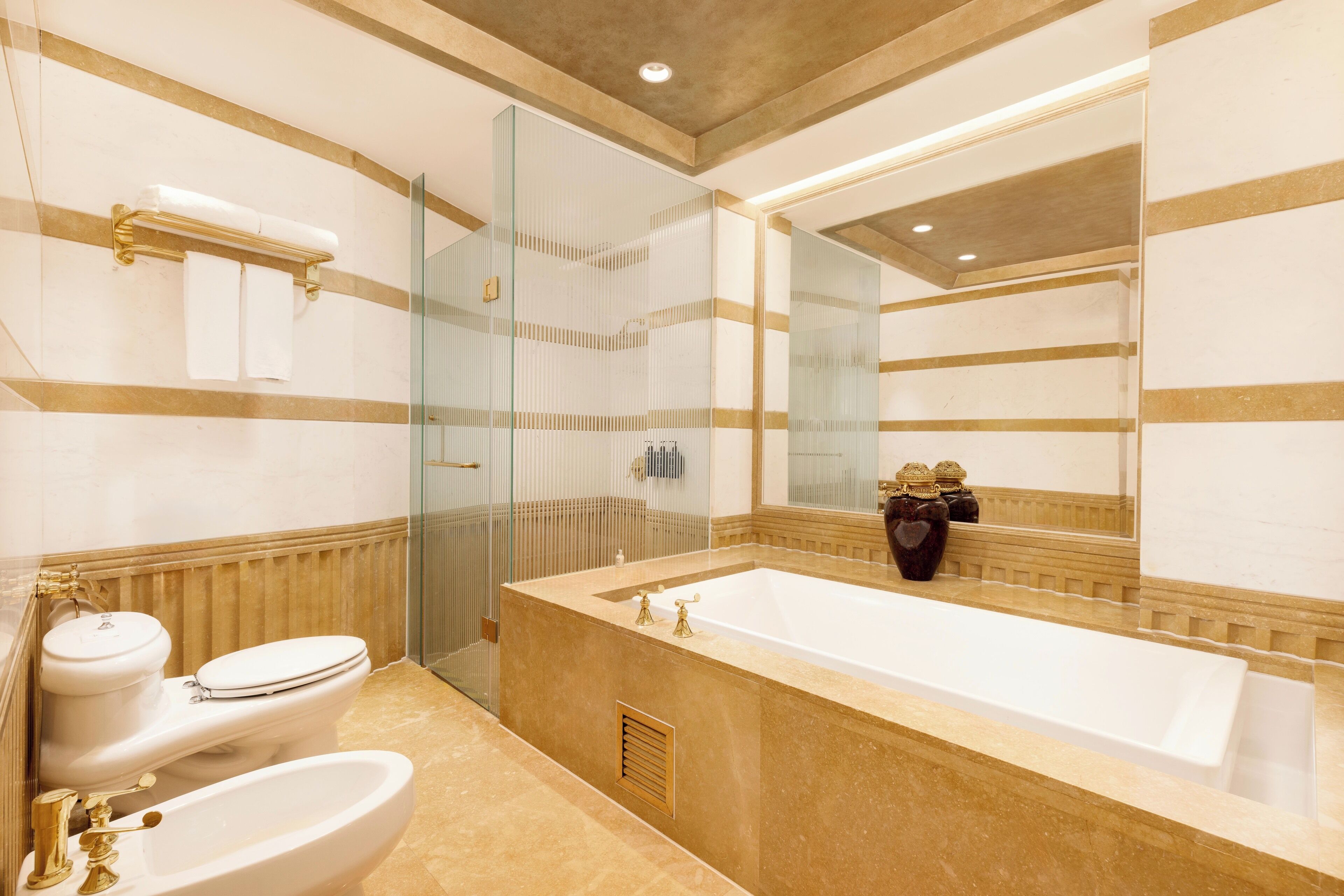 A bathroom with a large bathtub, two toilets, a glass shower enclosure, and a mirror with a decorative vase.