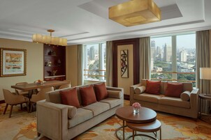 A spacious living room with a sofa, a round coffee table, and a dining area with chairs and a table. Large windows offer a city view.