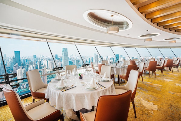 Un espace repas avec des tables rondes dressées pour le repas, de grandes fenêtres offrant une vue panoramique sur la ville et un design moderne au niveau du plafond.