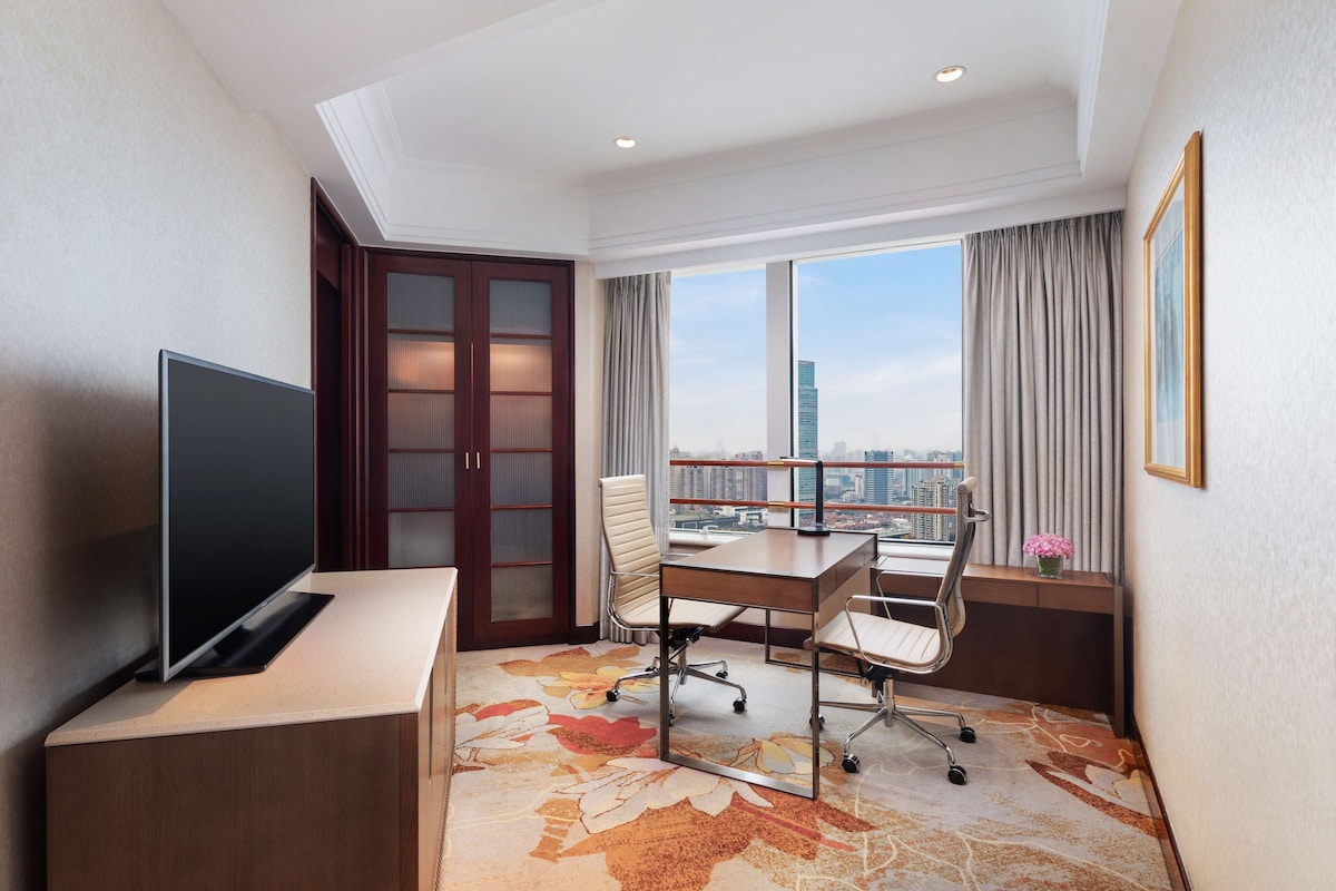 A hotel room with a large flat-screen TV, a wooden desk, two chairs, a large window with a city view, and a decorative carpet.