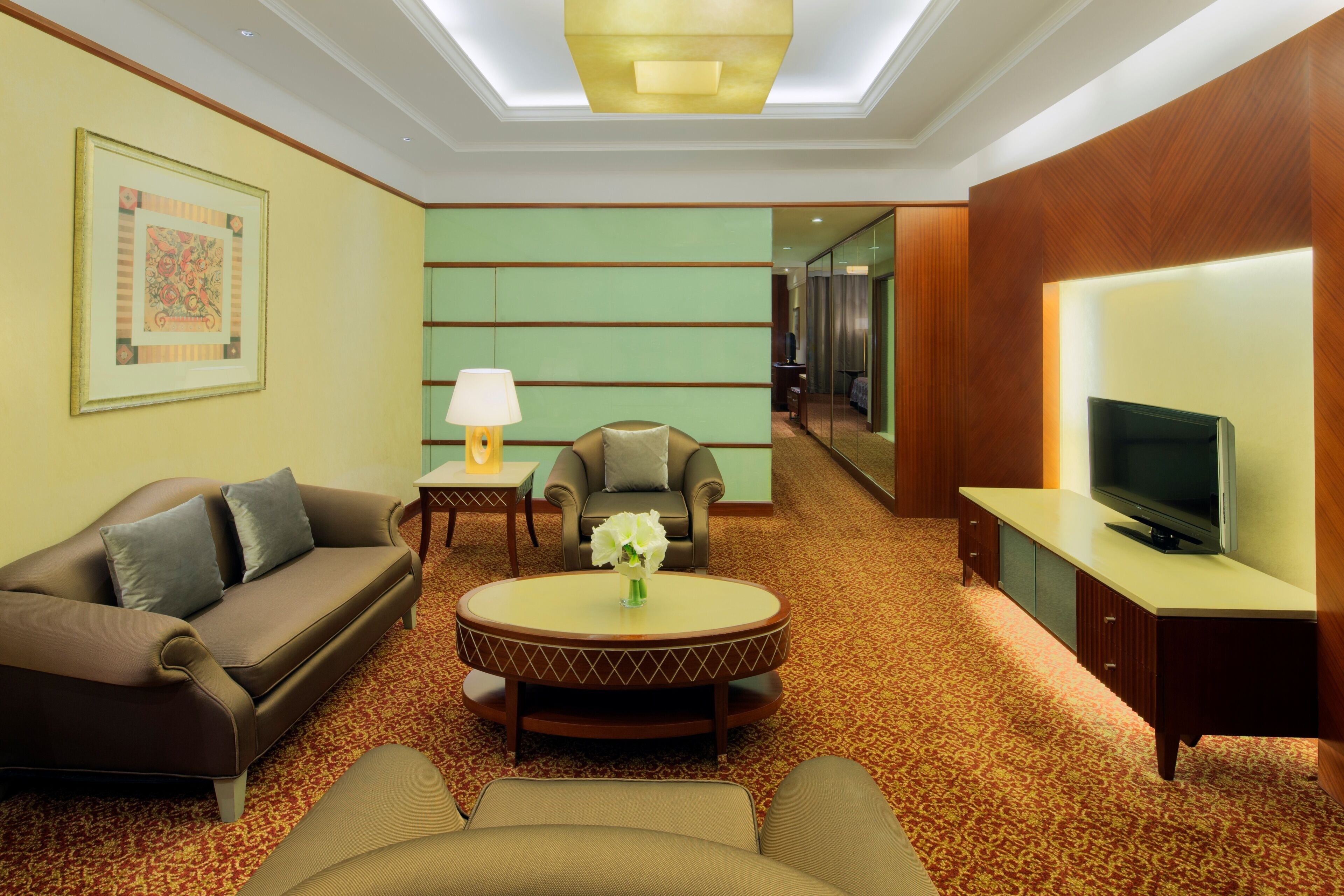 Una habitación de hotel con un sofá marrón, una mesa redonda con un florero, un televisor sobre un mueble de madera y un espejo en la pared.
