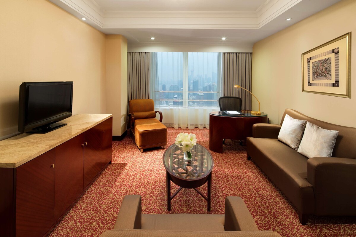 A hotel room with a TV, sofa, armchair, and a small table with a vase.