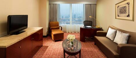 A hotel room with a TV, sofa, armchair, and a small table with a vase.