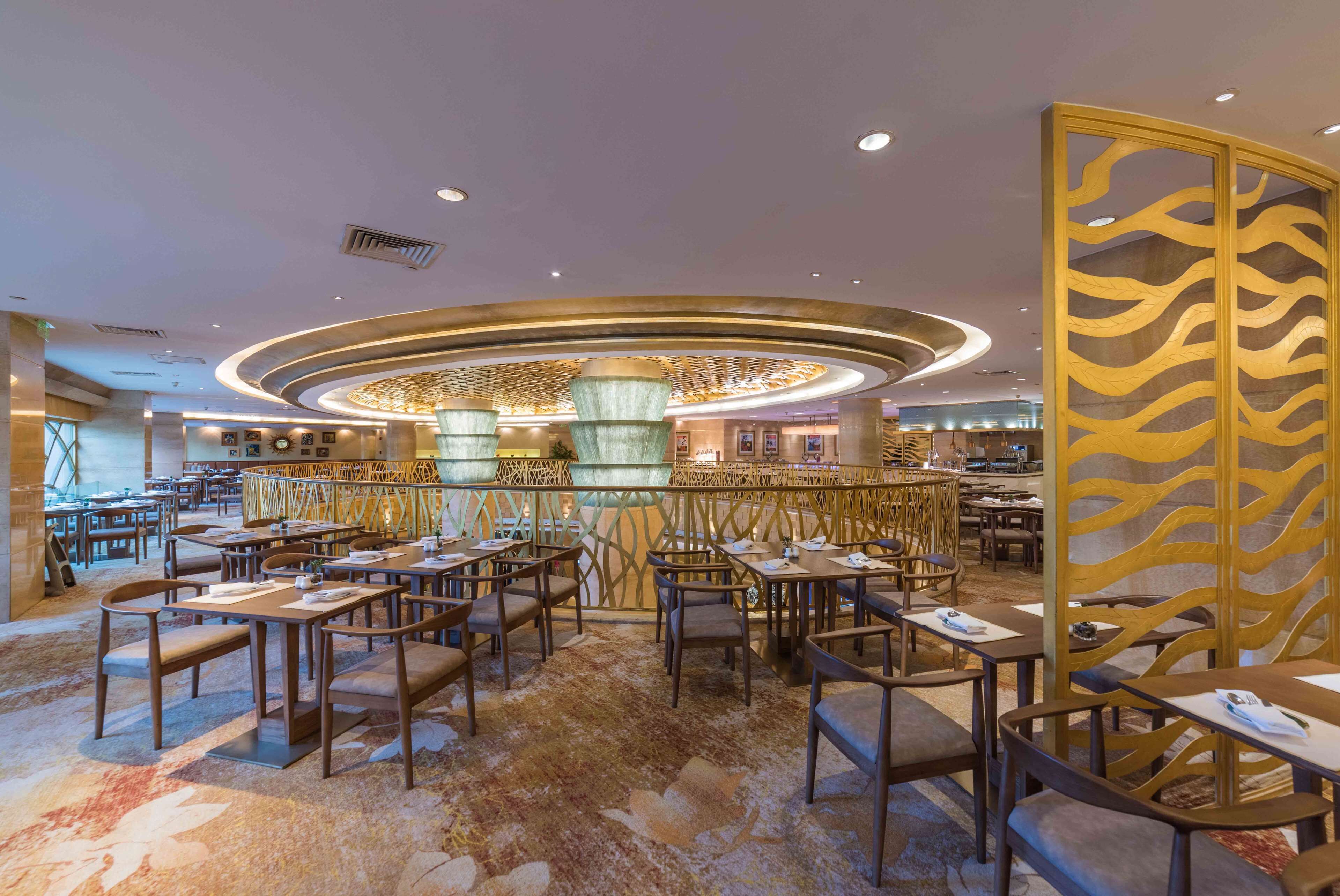 En rummelig restaurant med et centralt, rundt lysarmatur, flere siddeområder og en dekorativ rumdeler med bladmotiv.