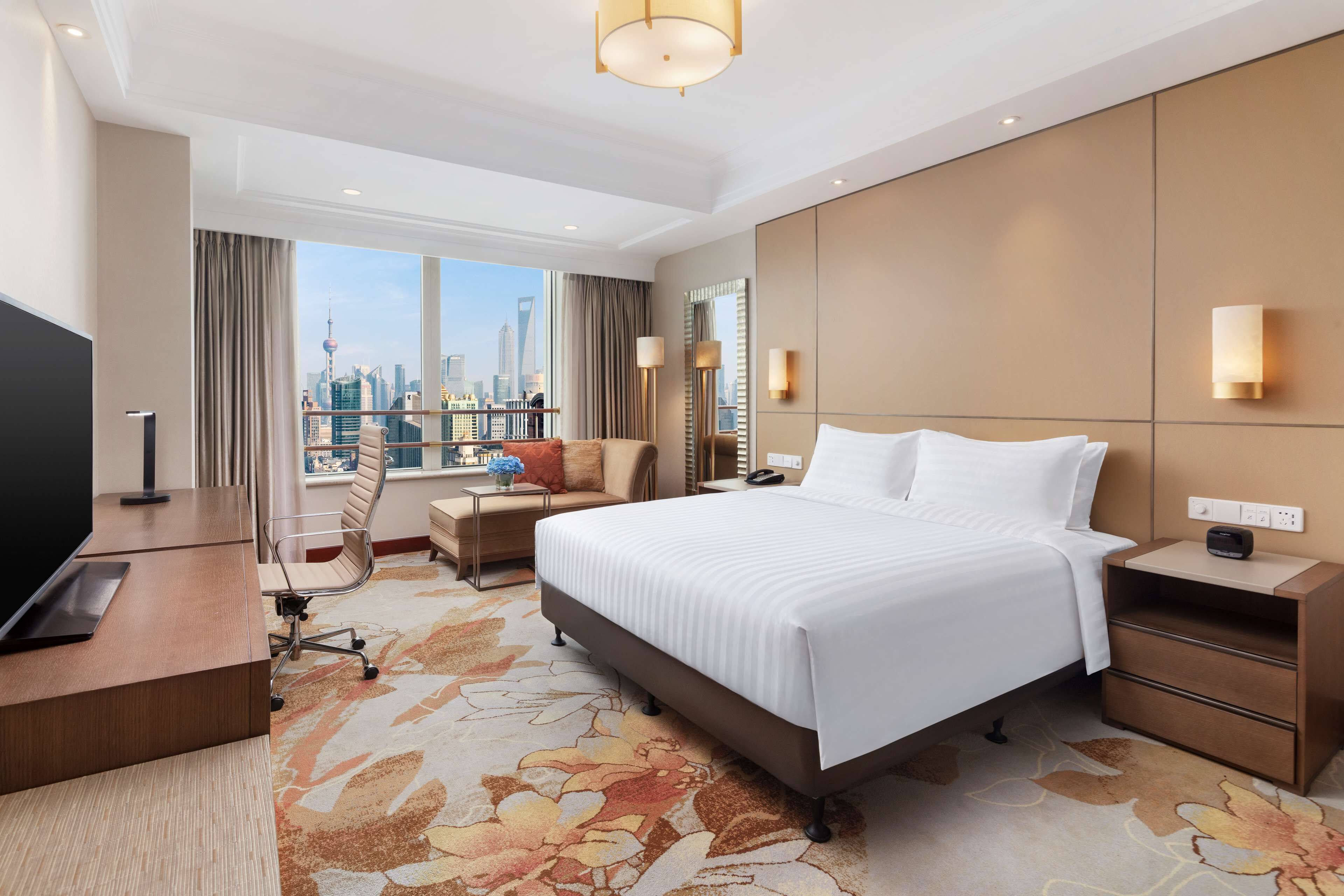 A hotel room with a large bed, a desk with a computer, a chair, and a view of a city skyline.