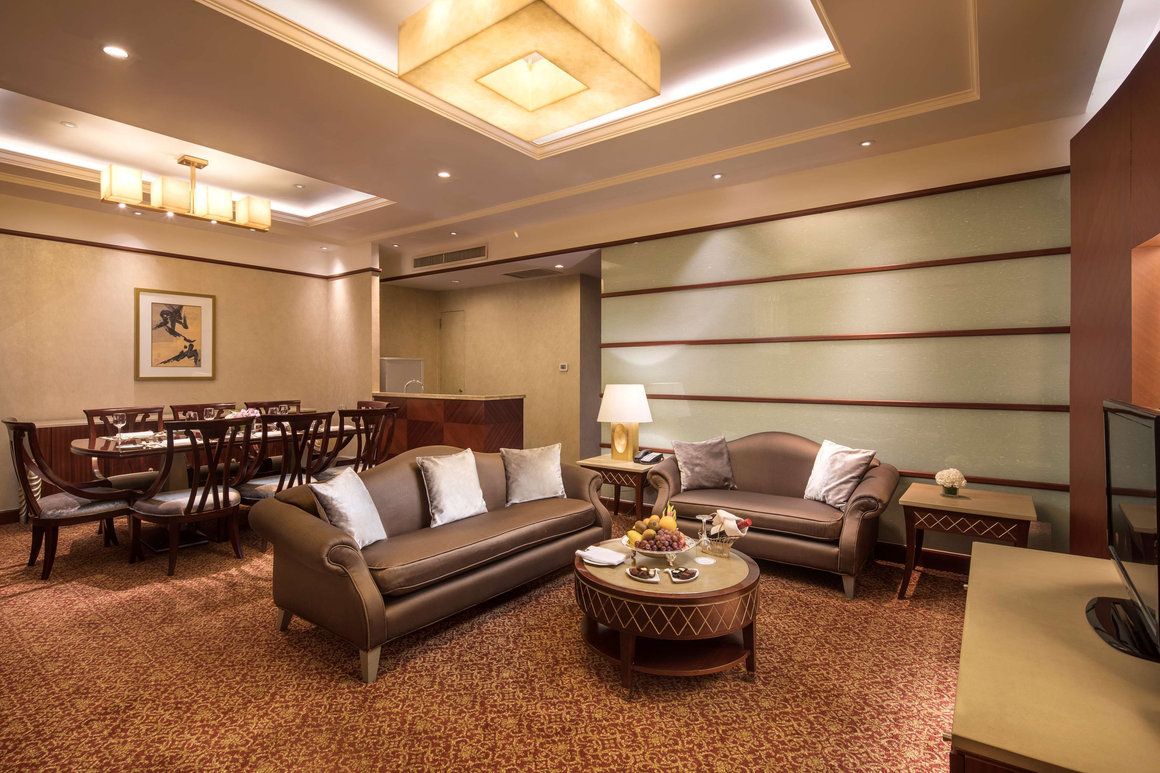 A hotel room with a brown sofa, a dining table with chairs, a coffee table with fruit, and a TV on a stand.
