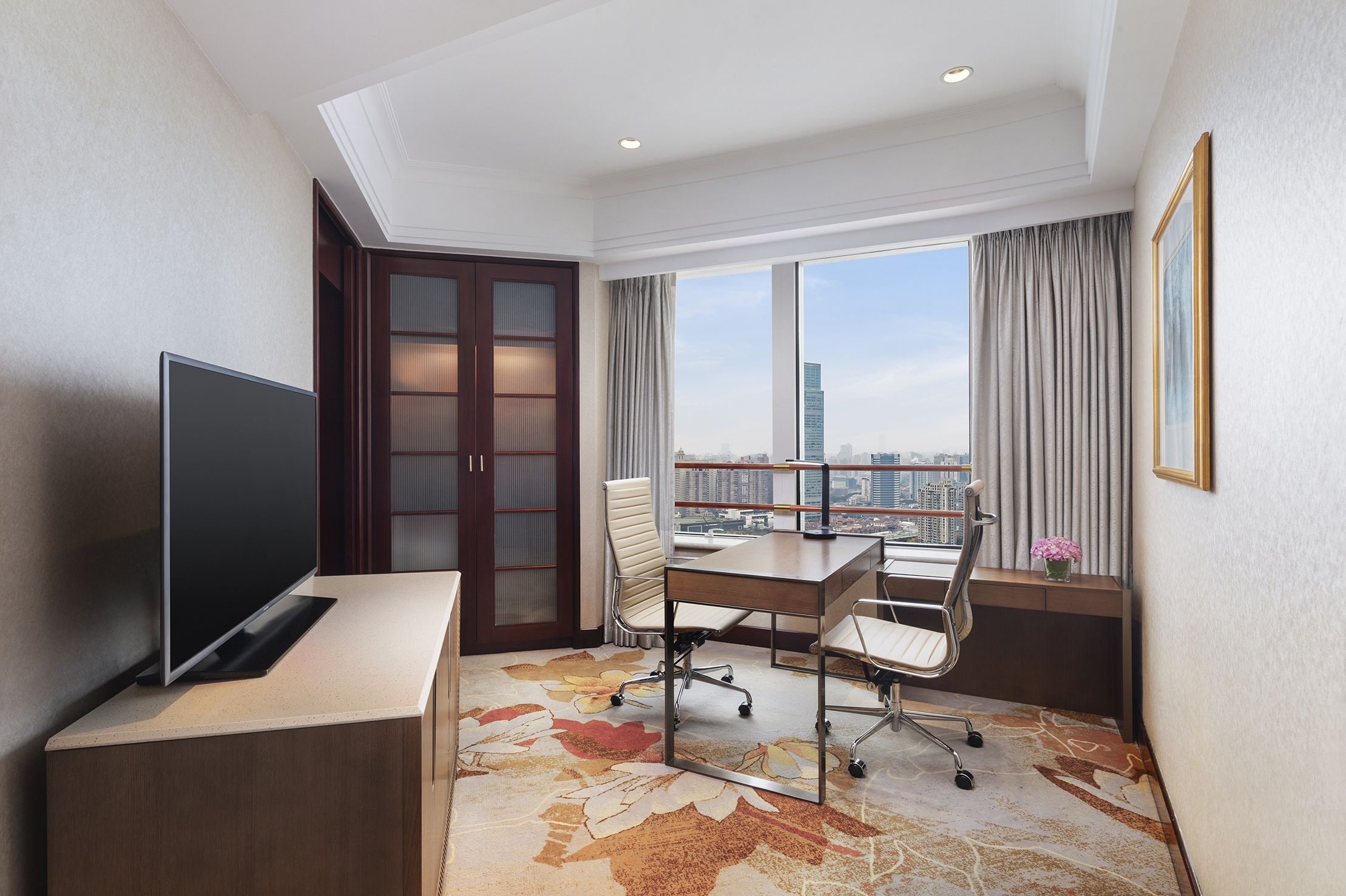 Quarto de hotel com escrivaninha, cadeiras, televisão, janela grande com cortinas e vista para a paisagem urbana.