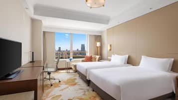 A hotel room with two beds, a desk with a TV, a small table, and a large window with a city view.