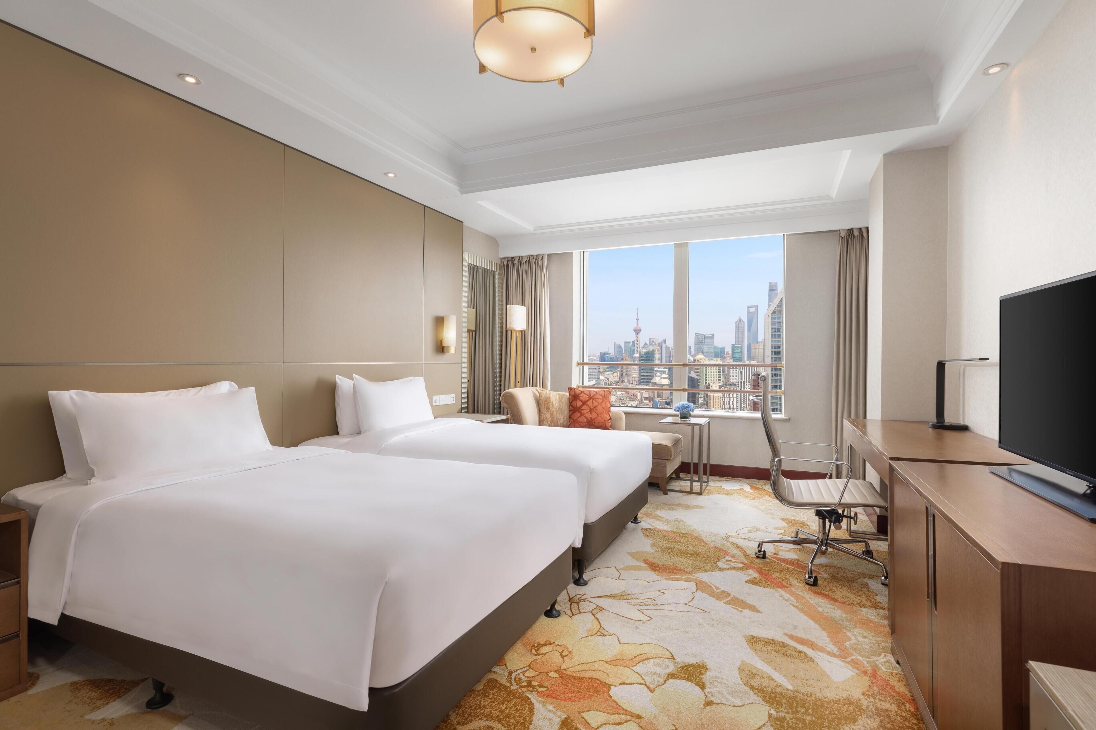 A hotel room with two beds, a desk, a TV, and a view of a cityscape.