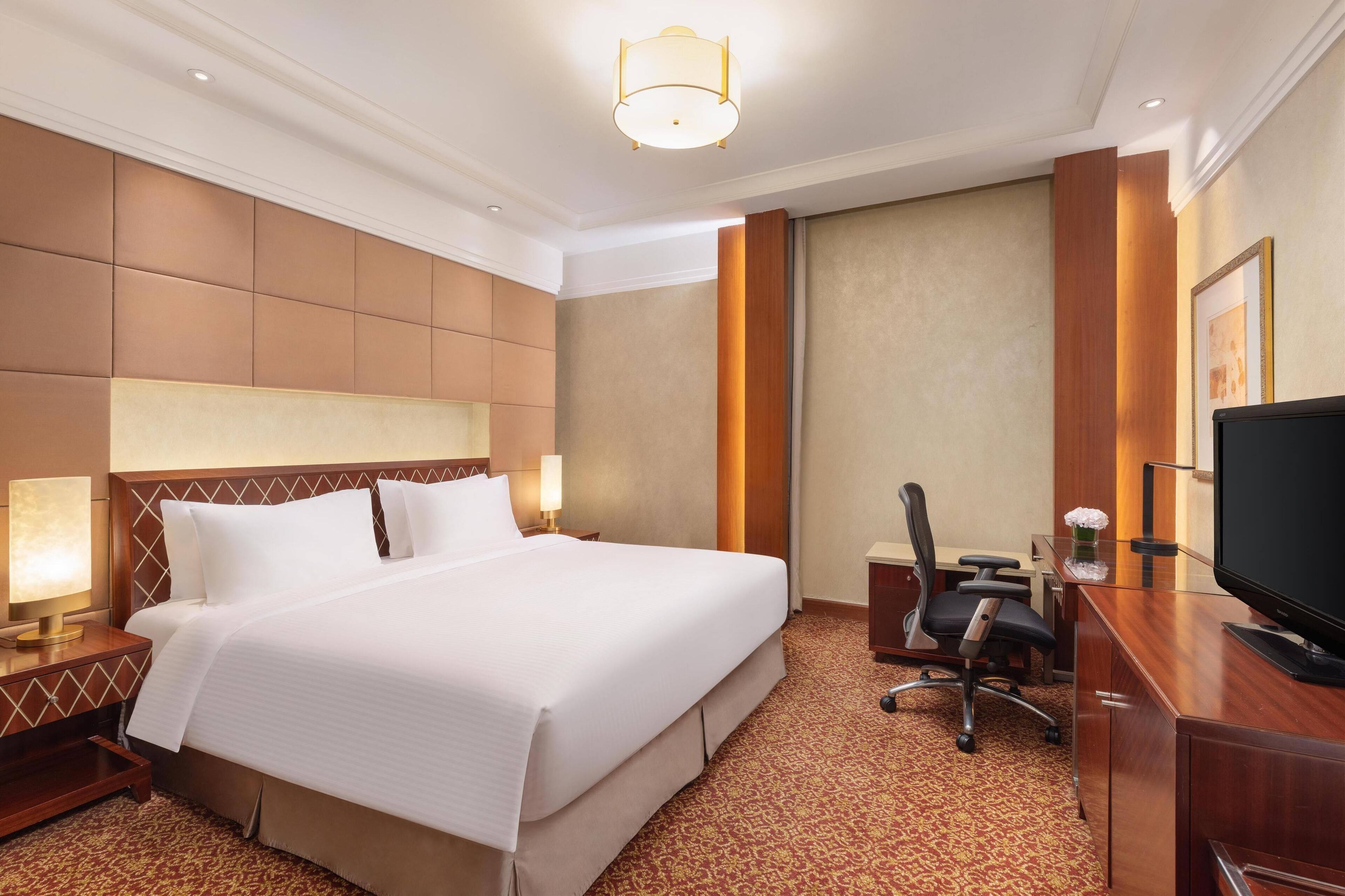 A hotel room with a large bed, a desk with a computer, a TV, and a lamp.