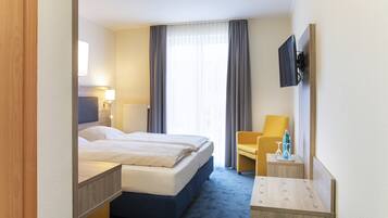 A hotel room with a double bed, a bedside table with a lamp, a chair, a small table with a bottle and glass, and a mounted TV.