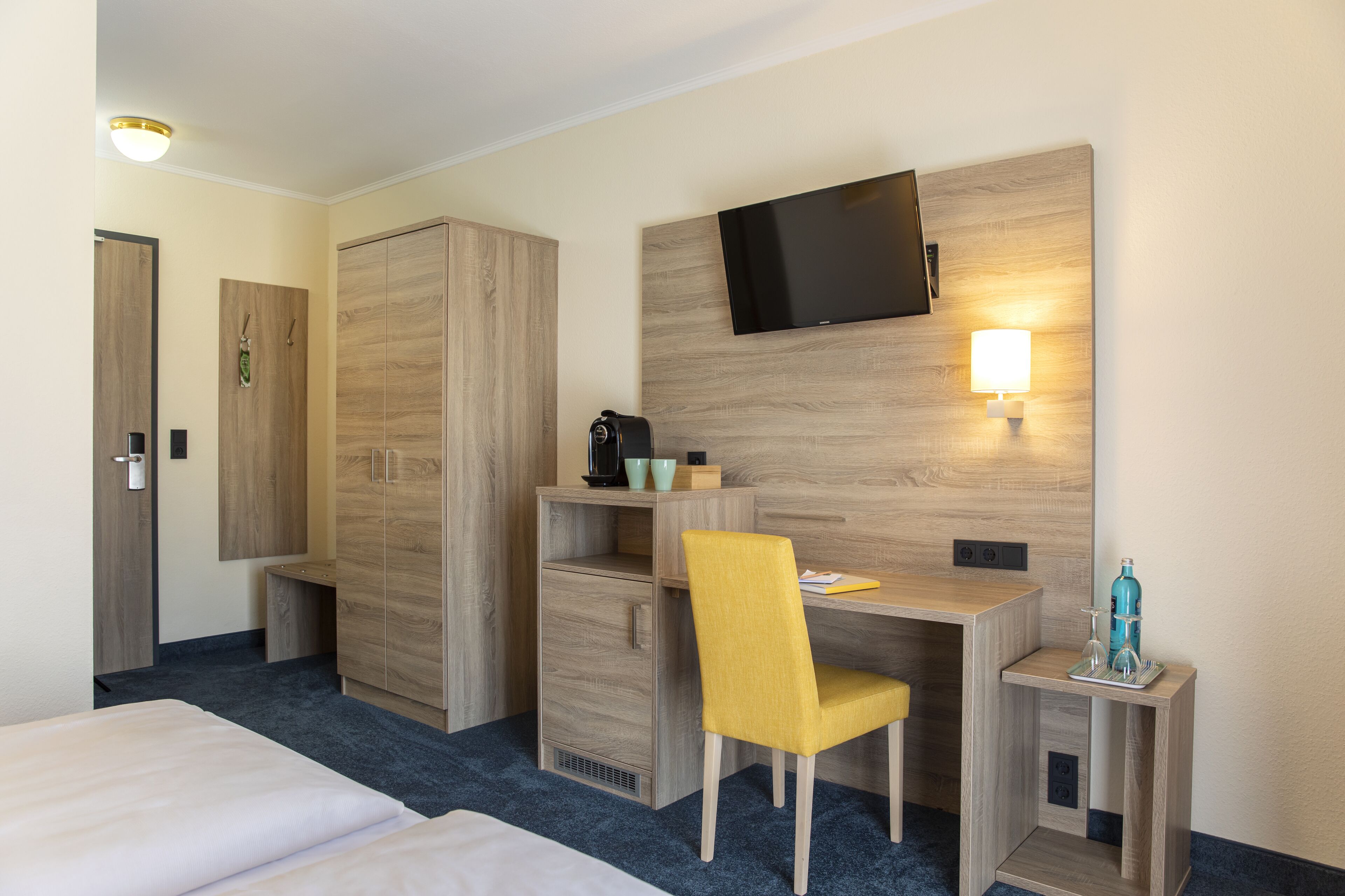 Ein Hotelzimmer mit einem Bett, einem Schreibtisch mit Stuhl, einem an der Wand befestigten Fernseher und einem kleinen Tisch mit Flasche und Gläsern.