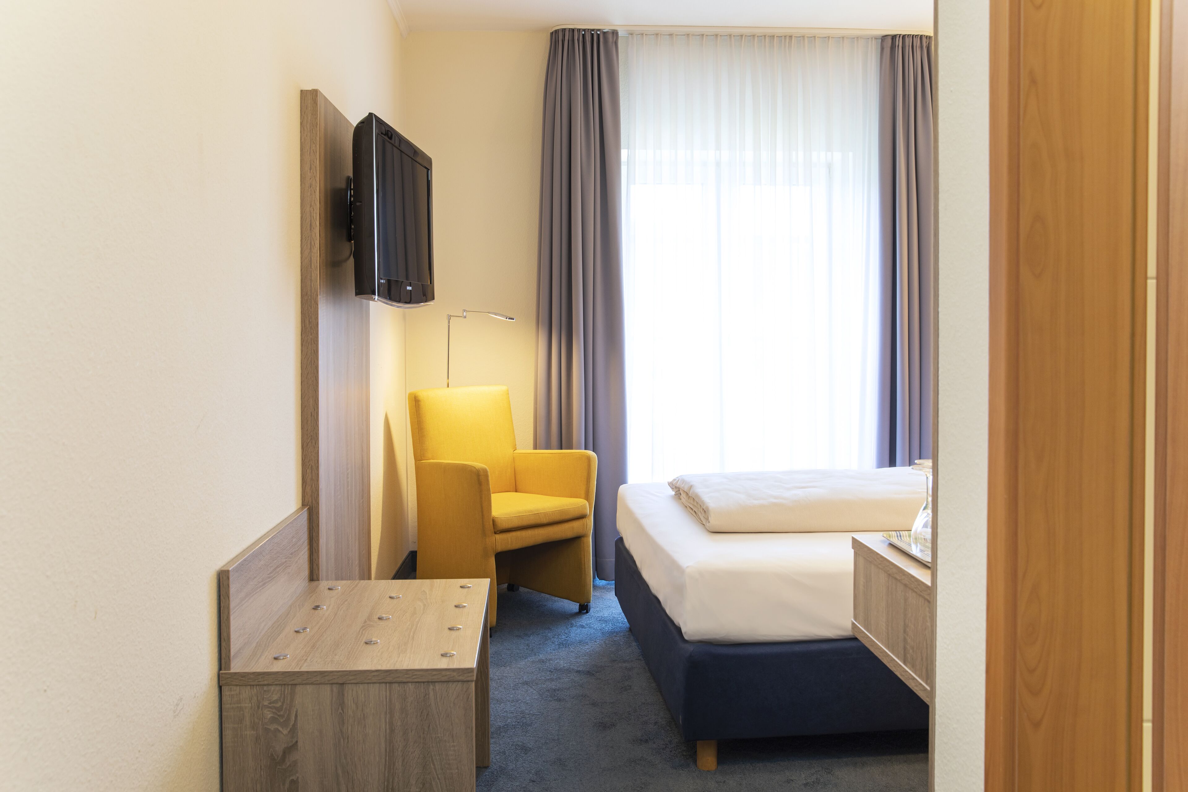 Een hotelkamer met een bed, een bureau, een stoel, een aan de muur bevestigde televisie en een raam met gordijnen.