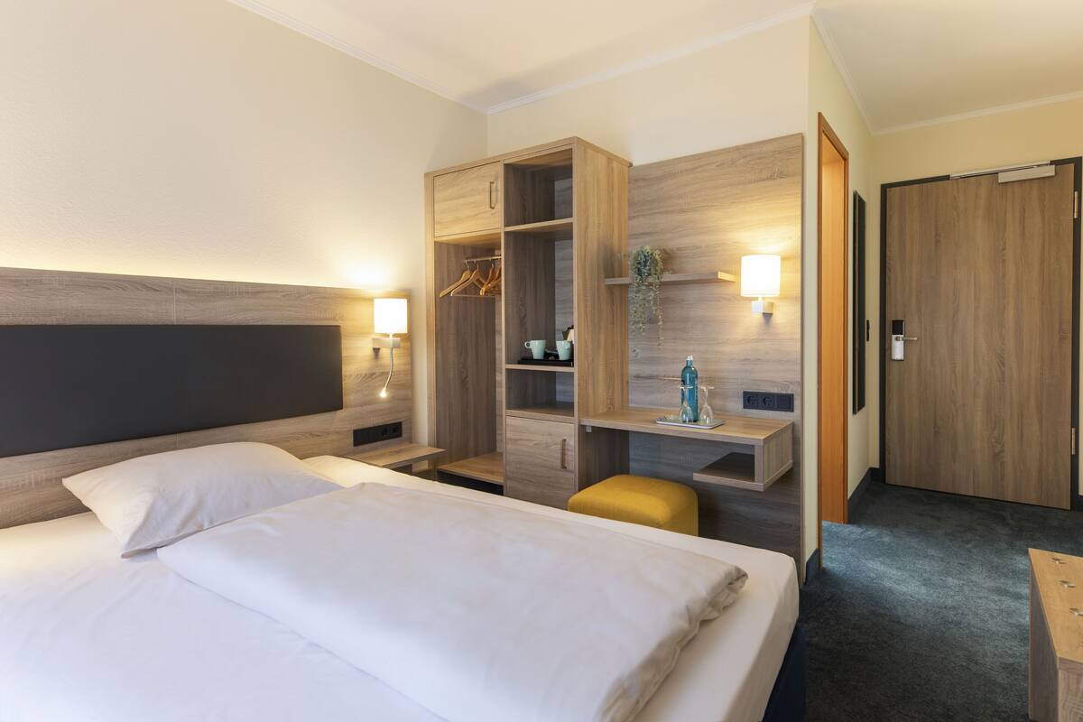 A hotel room with a large bed, a wooden wardrobe, a desk with a chair, and a small seating area.