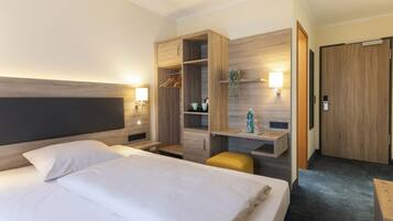 A hotel room with a large bed, a wooden wardrobe, a desk with a chair, and a small seating area.