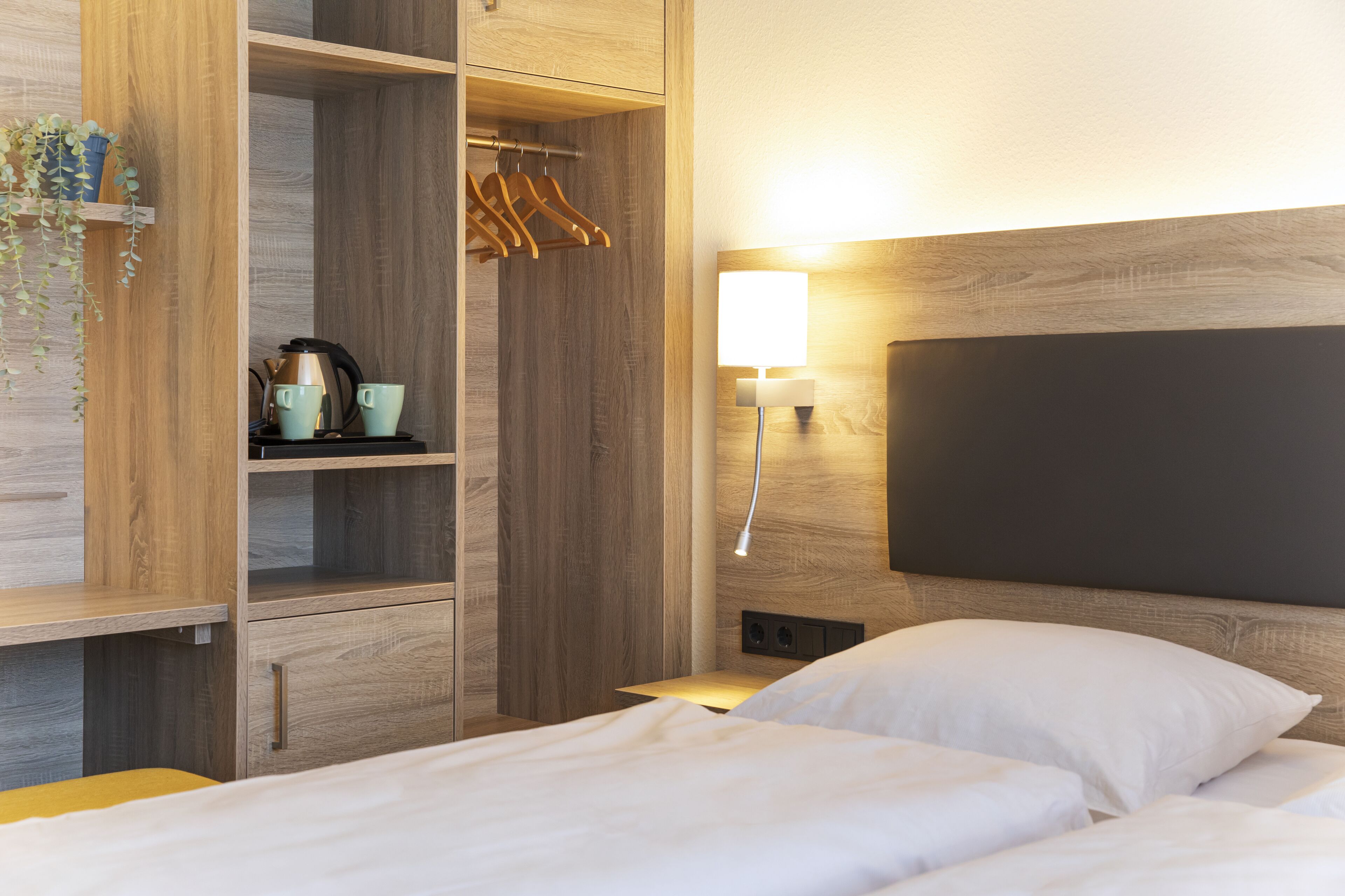 A modern bedroom with a wooden wardrobe, a bedside table with a lamp, and a bed with white bedding.