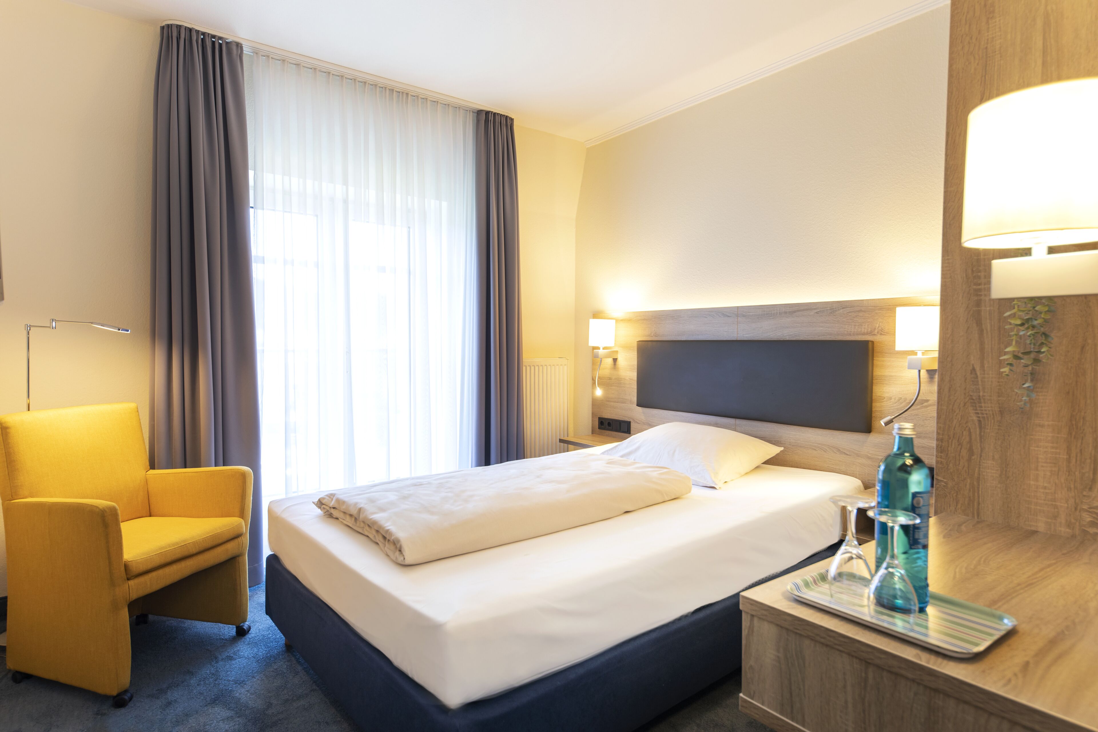 Habitación de hotel con cama, una silla, mesita de noche con una botella y vasos, y una lámpara.