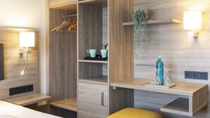 Une chambre d’hôtel moderne avec un lambris en bois, un lit, une table de chevet, une commode, une armoire, une étagère avec une plante et une lampe fixée au mur.