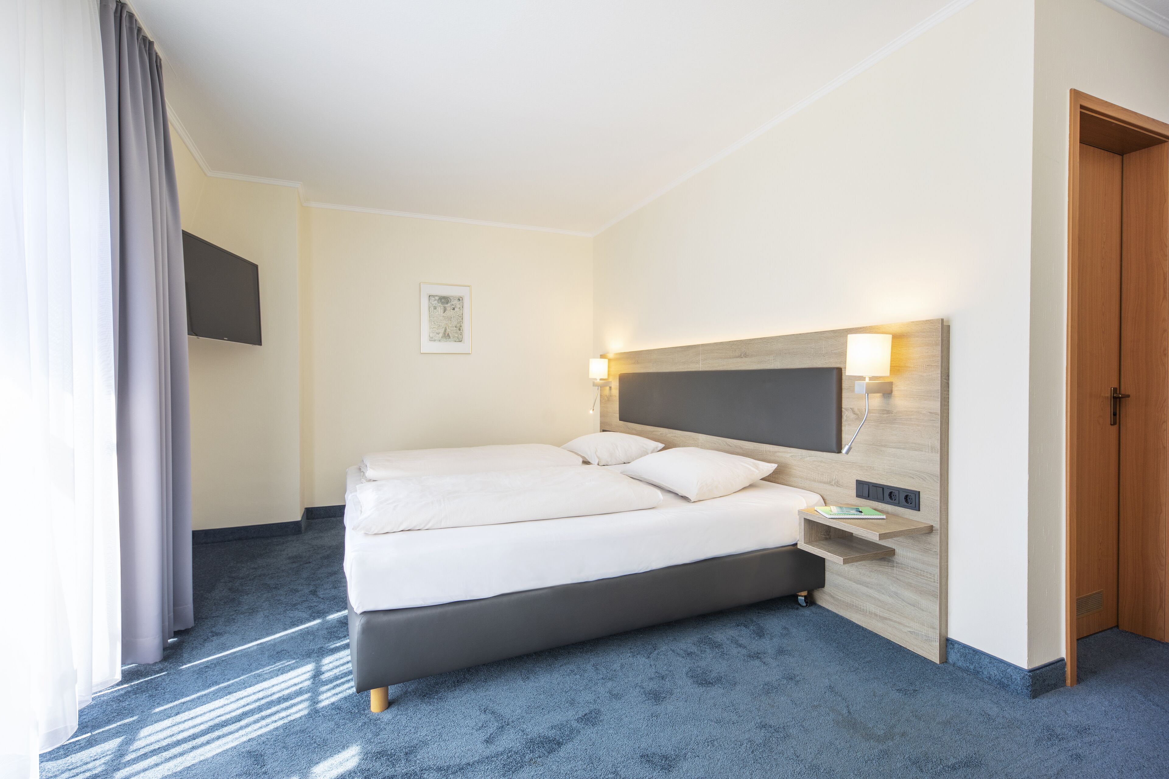 Habitación de hotel con una cama grande, un televisor de pantalla plana montado en la pared, una mesita de noche con lámpara y una puerta de madera.