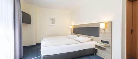 Une chambre d’hôtel avec un grand lit, un téléviseur à écran plat fixé au mur, une table de chevet avec une lampe et une porte en bois.