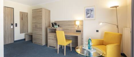 A modern hotel room with a wooden wardrobe, a desk with a chair, a glass table with a bottle, and a yellow armchair.