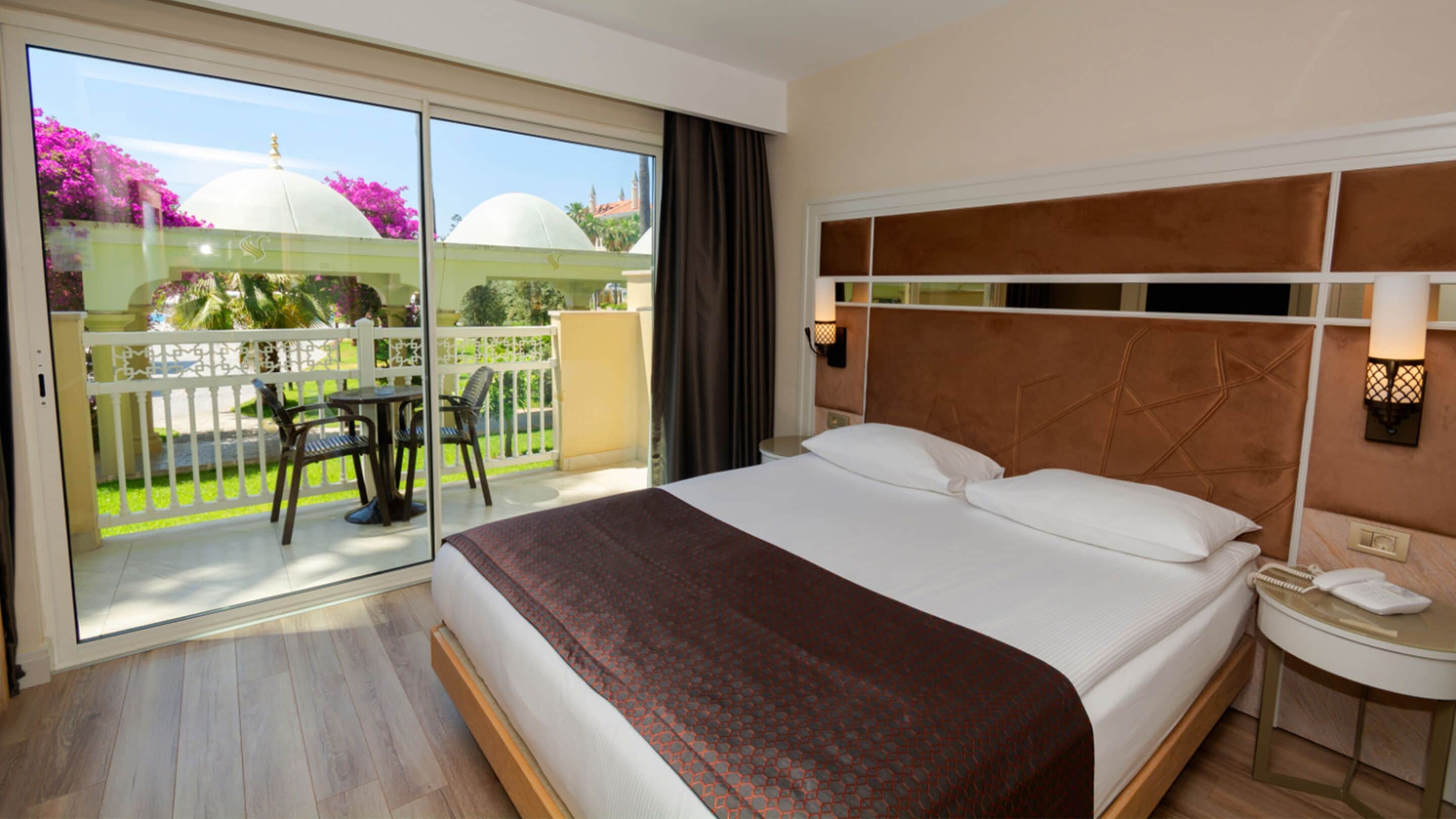Een hotelkamer met een groot bed, een nachtkastje, een telefoon en uitzicht op een balkon met tuinmeubilair en een tuin.