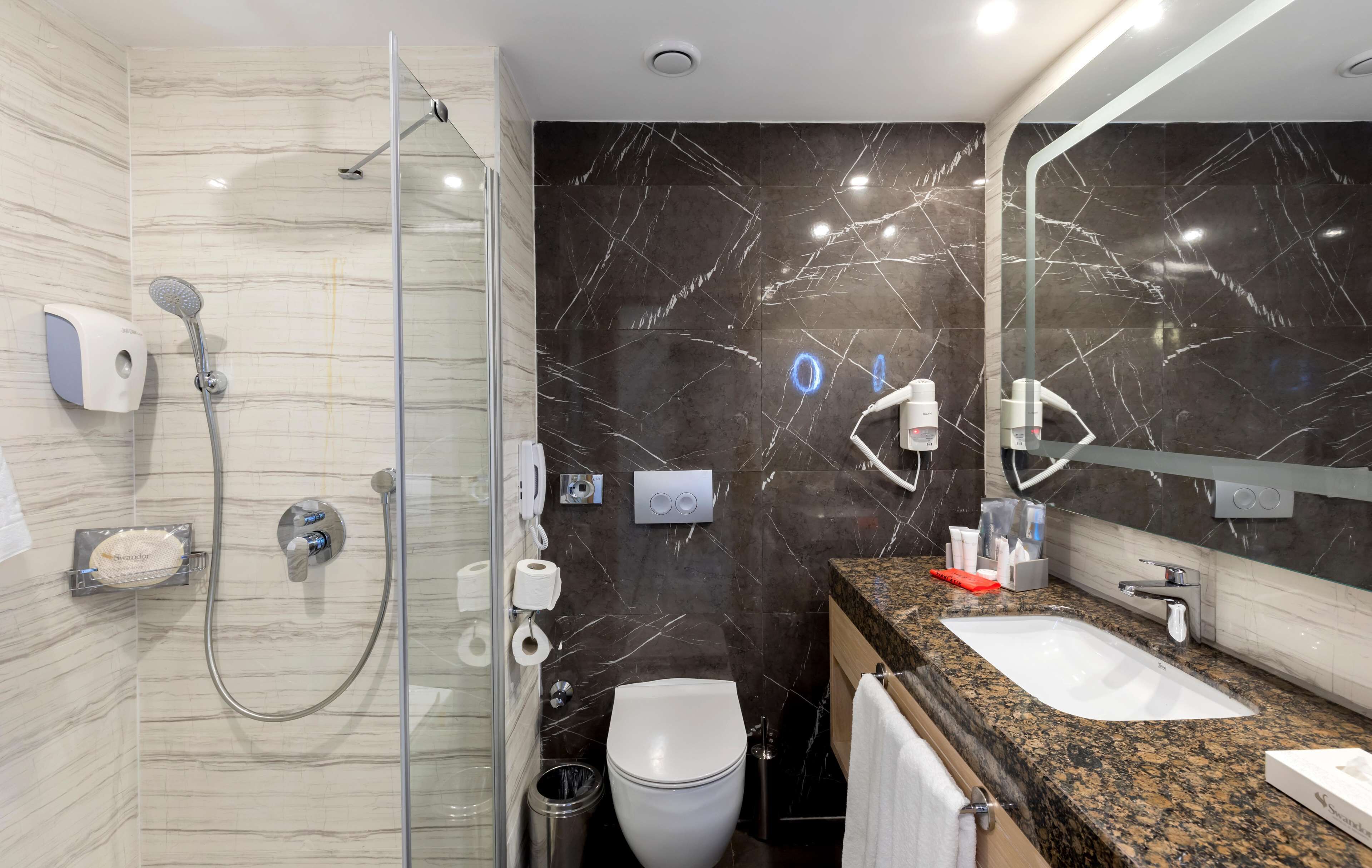 Une salle de bain moderne avec une douche à parois de verre, un mur recouvert de carreaux de marbre, un lavabo avec un plan de travail en granit et un miroir.
