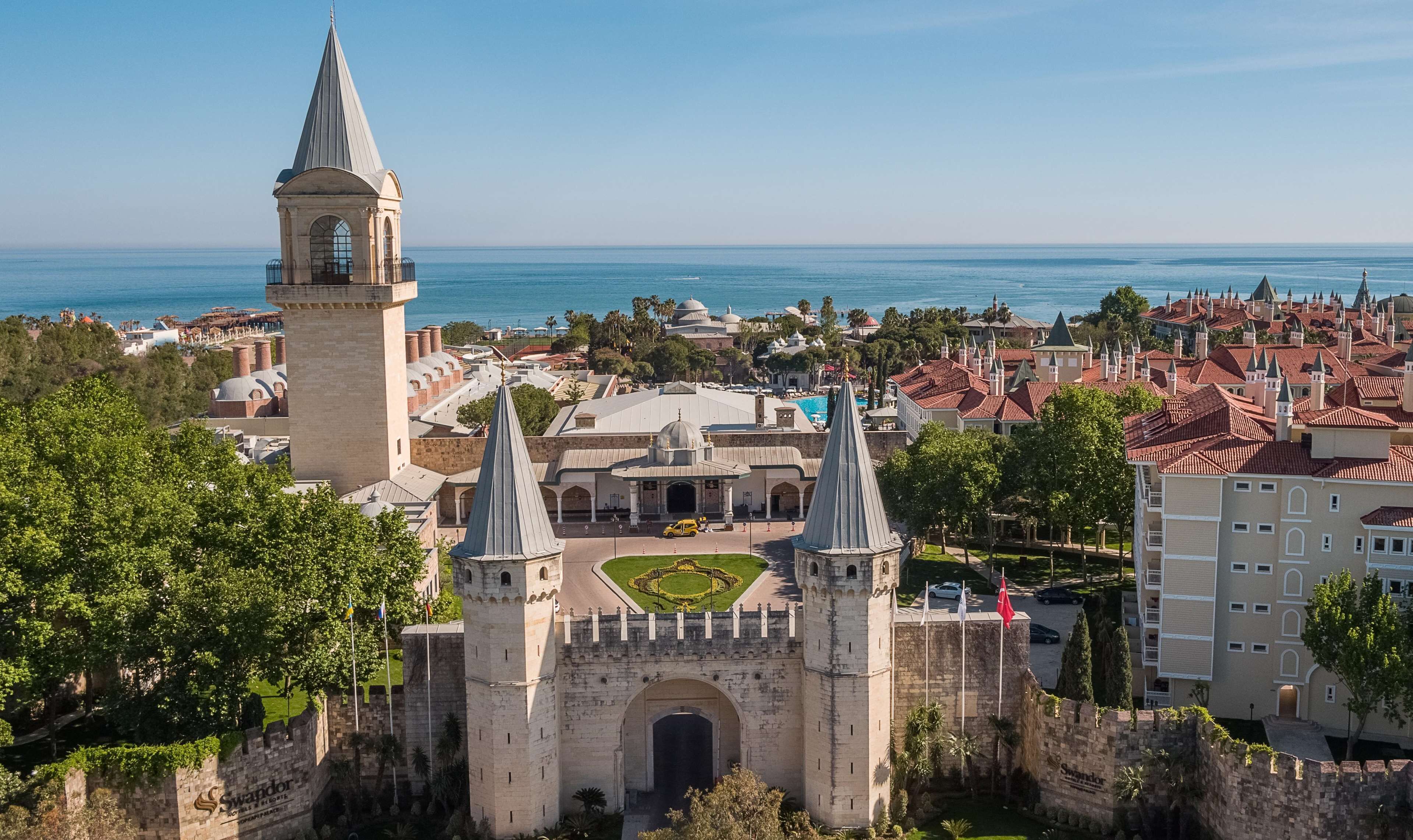 A historic castle with two prominent towers, a central courtyard, and a view of the sea.