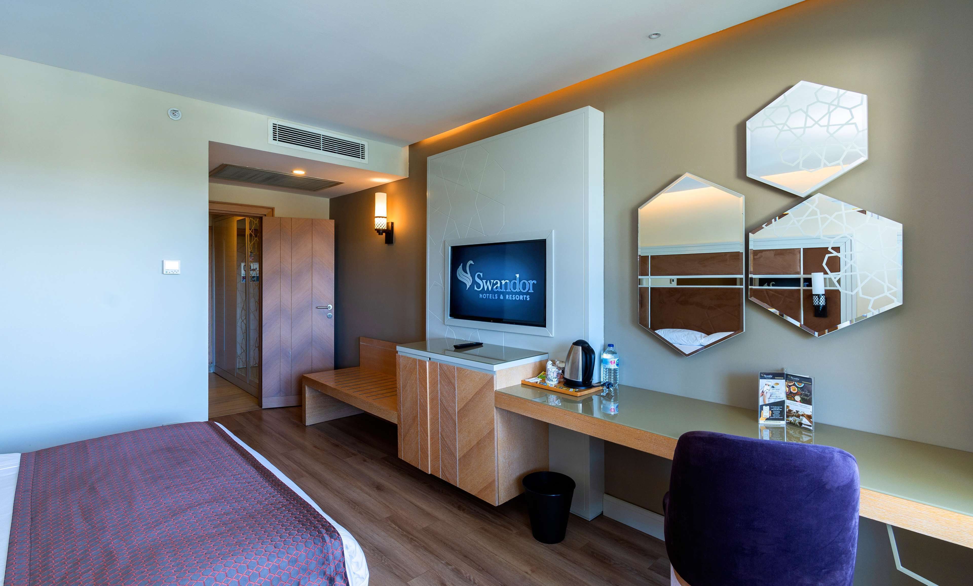 Quarto de hotel com uma cama, uma mesa com computador, uma televisão e um espelho fixado na parede.