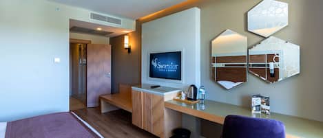 Quarto de hotel com uma cama, uma mesa com computador, uma televisão e um espelho fixado na parede.