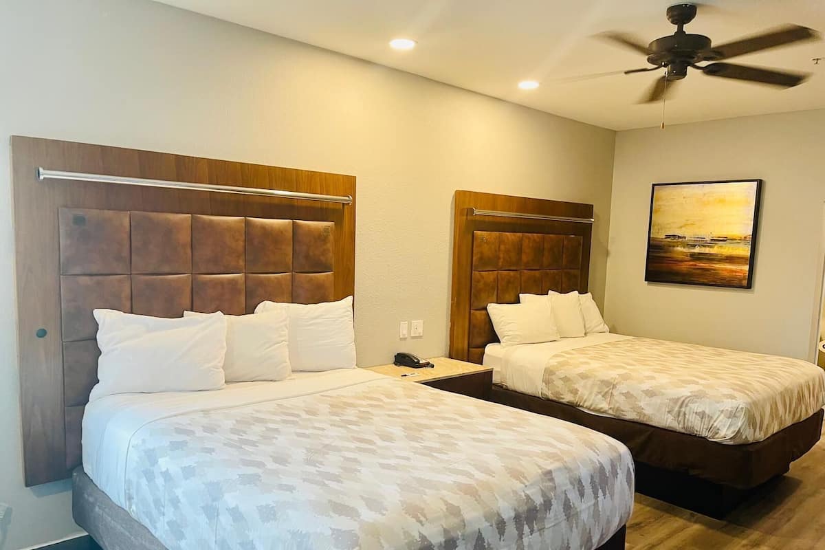 A hotel room with two beds, each with a wooden headboard and white bedding. There is a ceiling fan, a painting on the wall, and a bedside table with a phone.