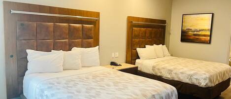 A hotel room with two beds, each with a wooden headboard and white bedding. There is a ceiling fan, a painting on the wall, and a bedside table with a phone.