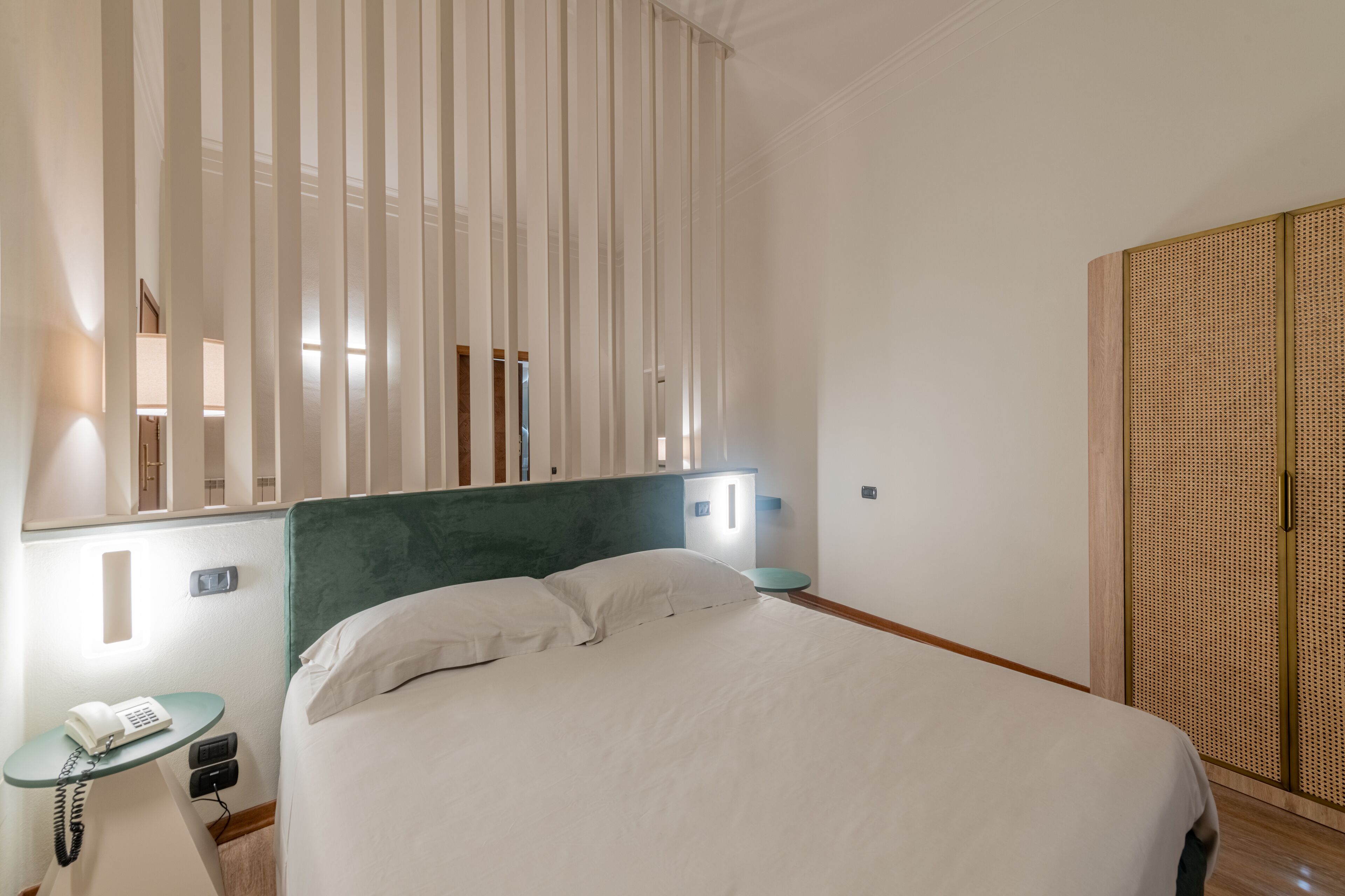Habitación de hotel con cama, mesita de noche con teléfono, luz de pared y armario con diseño tejido.