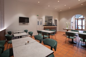 Un restaurante con sillas y mesas tapizadas de verde, un televisor de pantalla plana montado en la pared y una cocina visible a través de un arco.