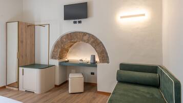 A modern hotel room with a stone fireplace, a flat-screen TV, a wooden wardrobe, a green sofa, and a small table with a kettle.