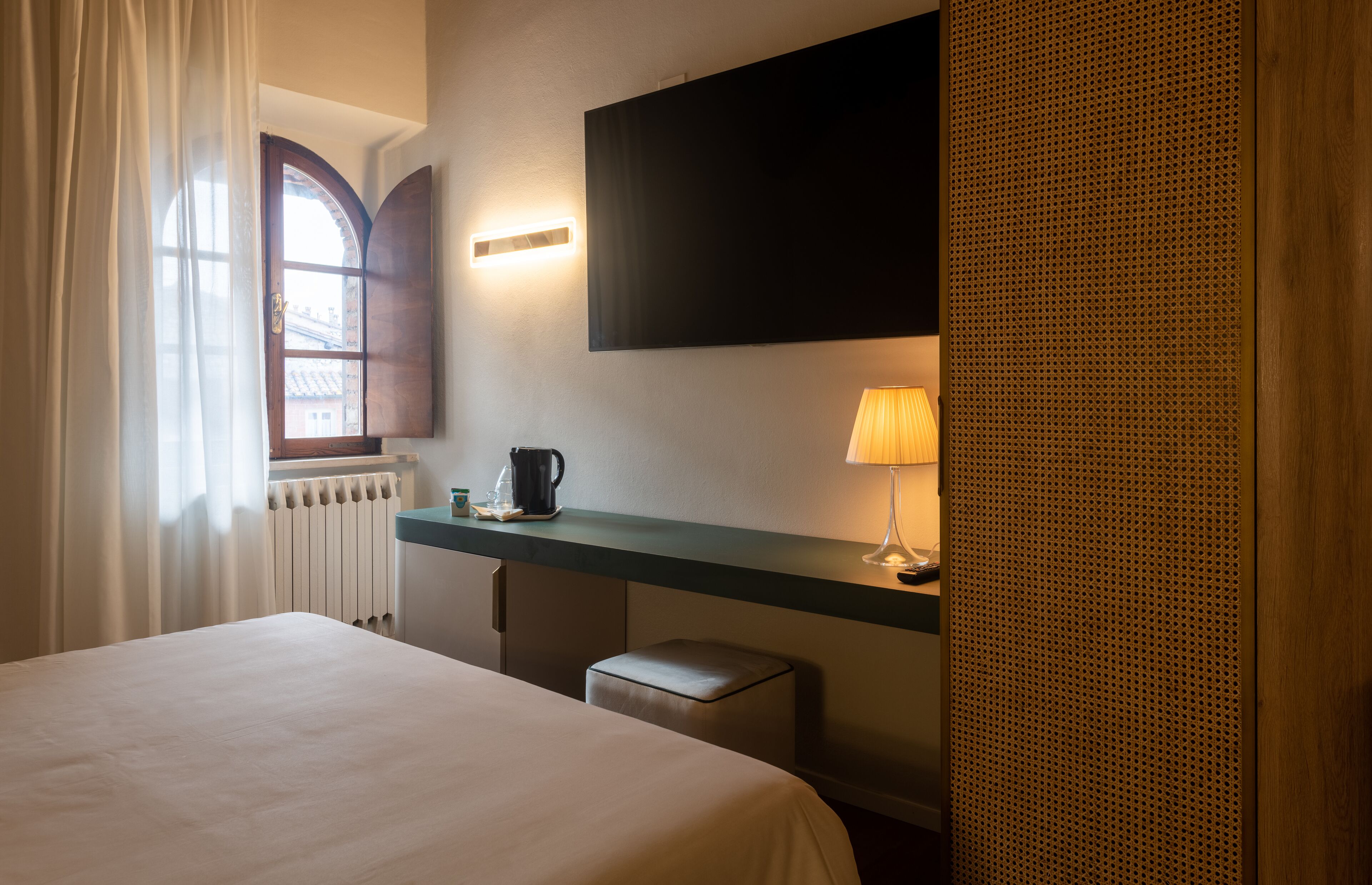 A hotel room with a bed, a desk with a lamp, a radiator, and a window with curtains.