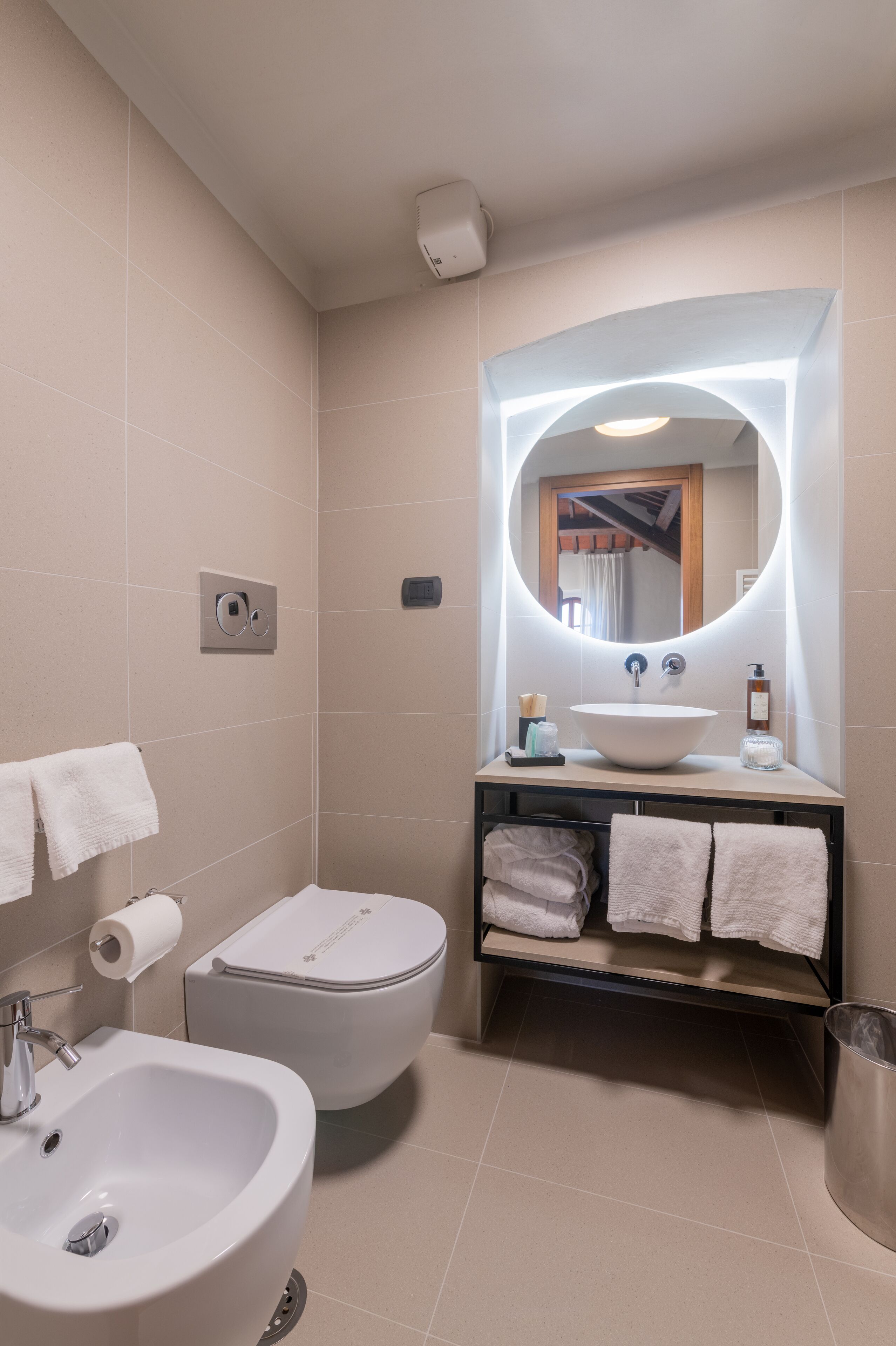 A modern bathroom with a white sink, a large mirror, a toilet, and a bidet. There are towels hanging on a rack and a trash can.