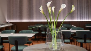 Una mesa con un florero transparente con flores blancas, ubicada en un comedor con sillas y mesas verdes.