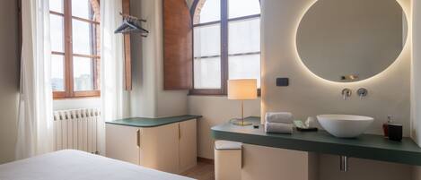 Una habitación de hotel con cama, escritorio con lámpara, espejo redondo y un ventanal grande.