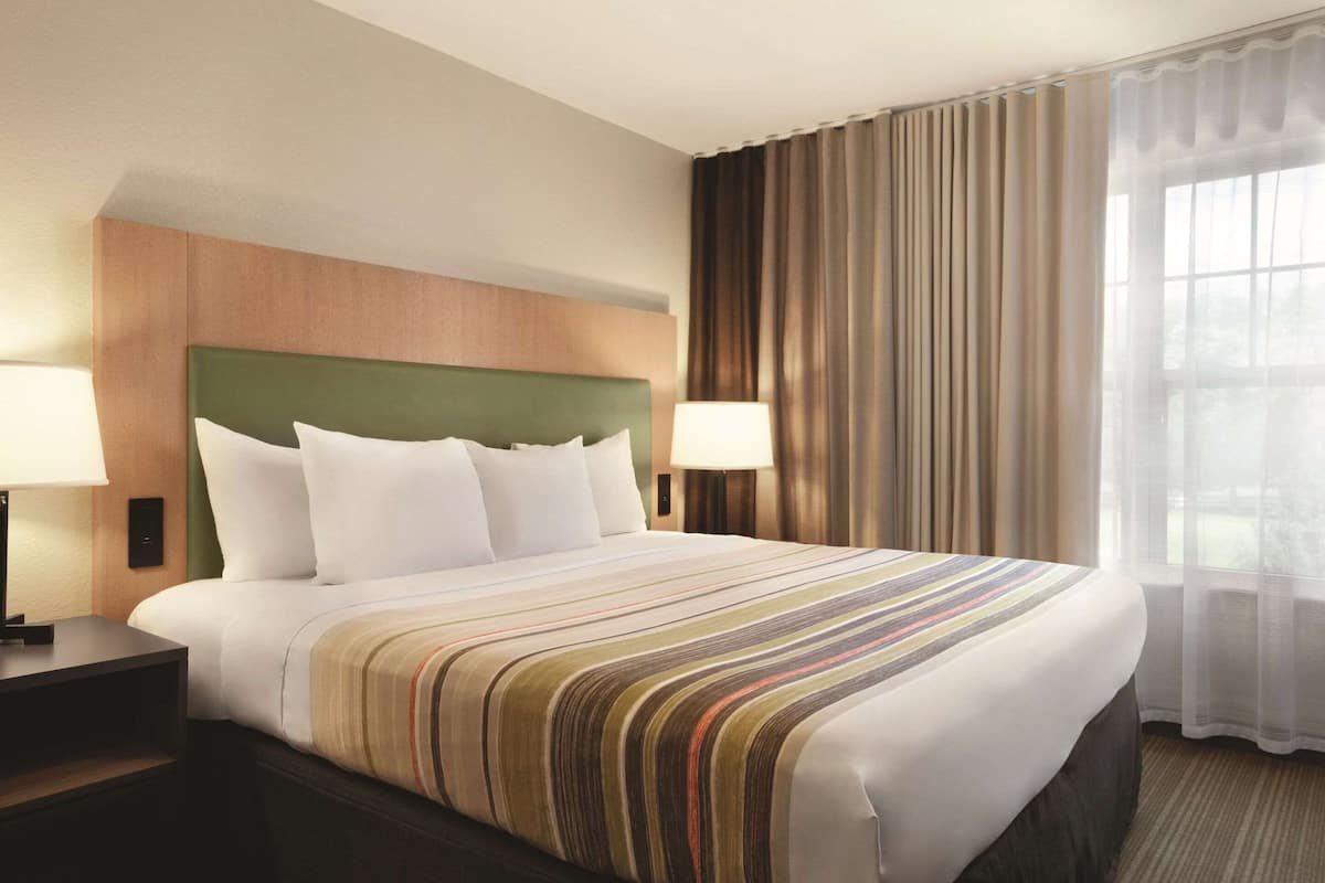 Quarto de hotel com uma cama grande, duas mesas de cabeceira, um espaldar verde e uma janela com cortinas.