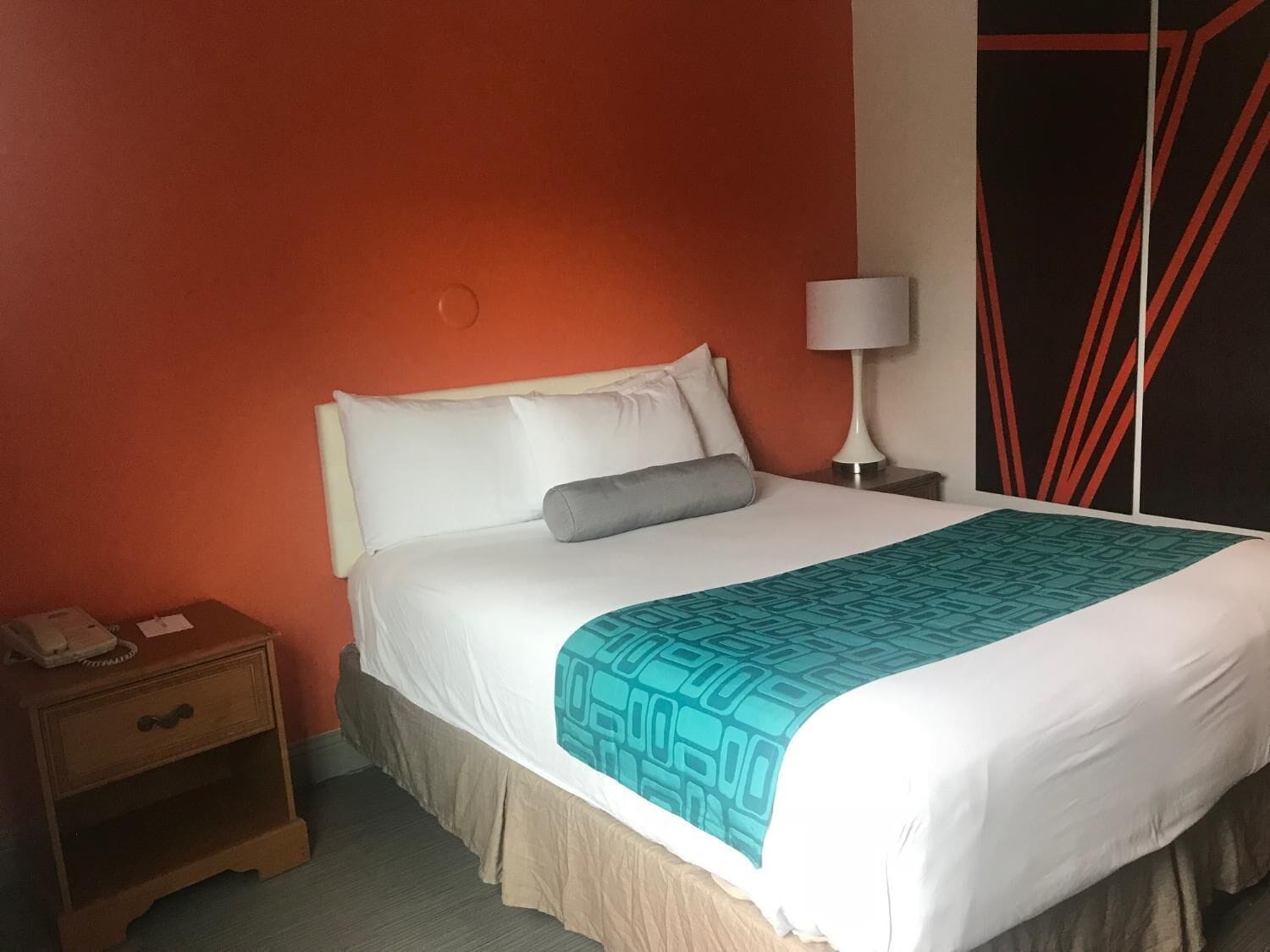 A neatly made bed with a white and teal bedspread, a grey pillow, and a white headboard in a room with orange walls and a black and red abstract wall art piece.