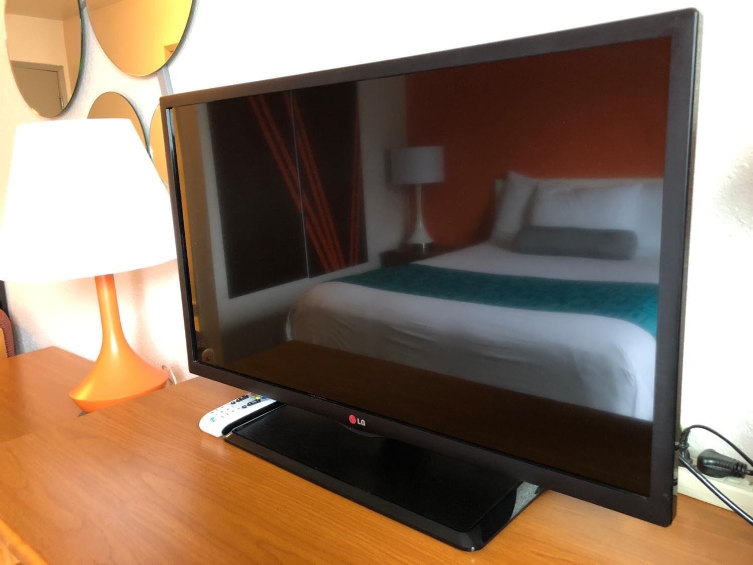 A hotel room with a large flat-screen TV on a wooden table, a remote control, and a bed with white bedding and a teal throw blanket.
