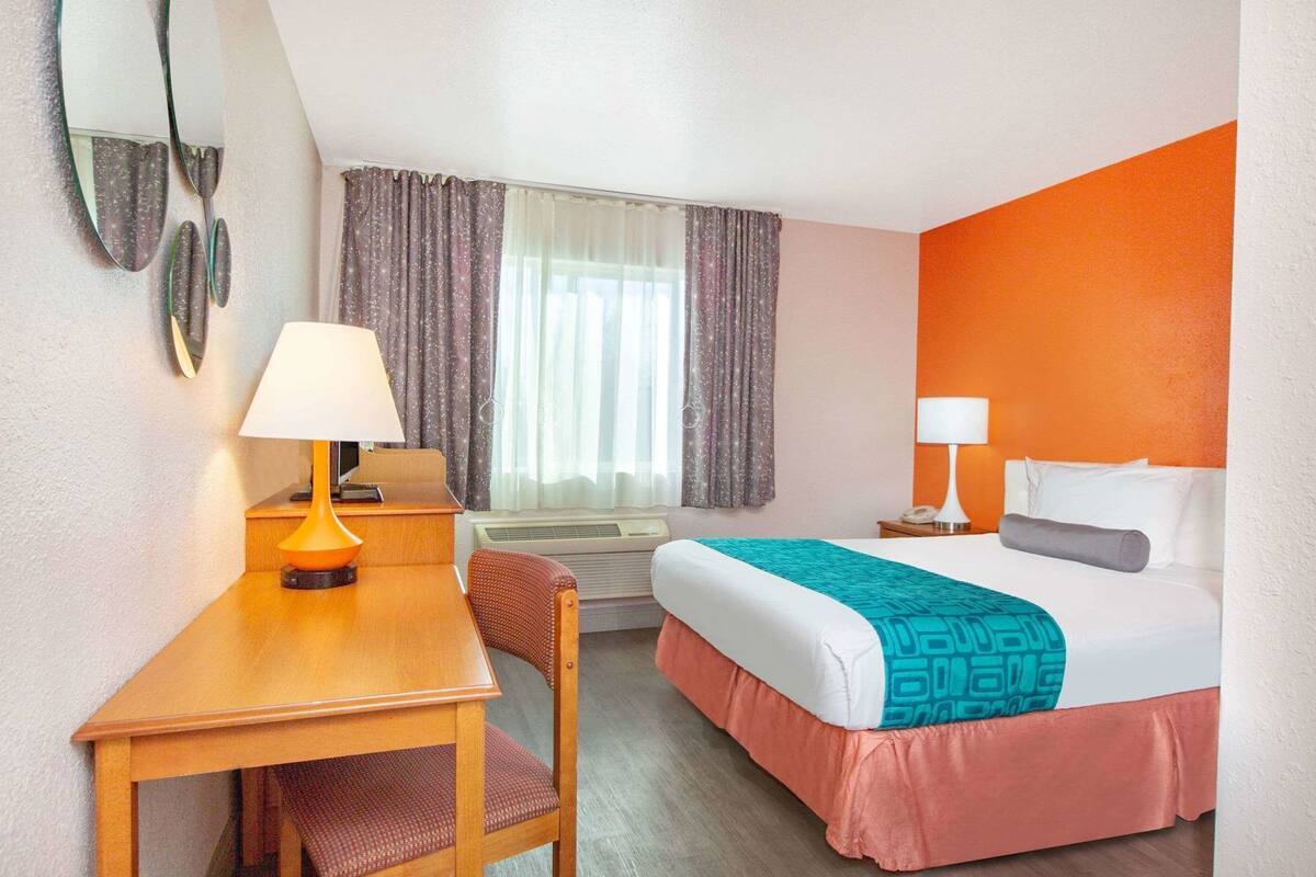 A hotel room with a bed, desk, chair, two lamps, a window with curtains, and an air conditioning unit.
