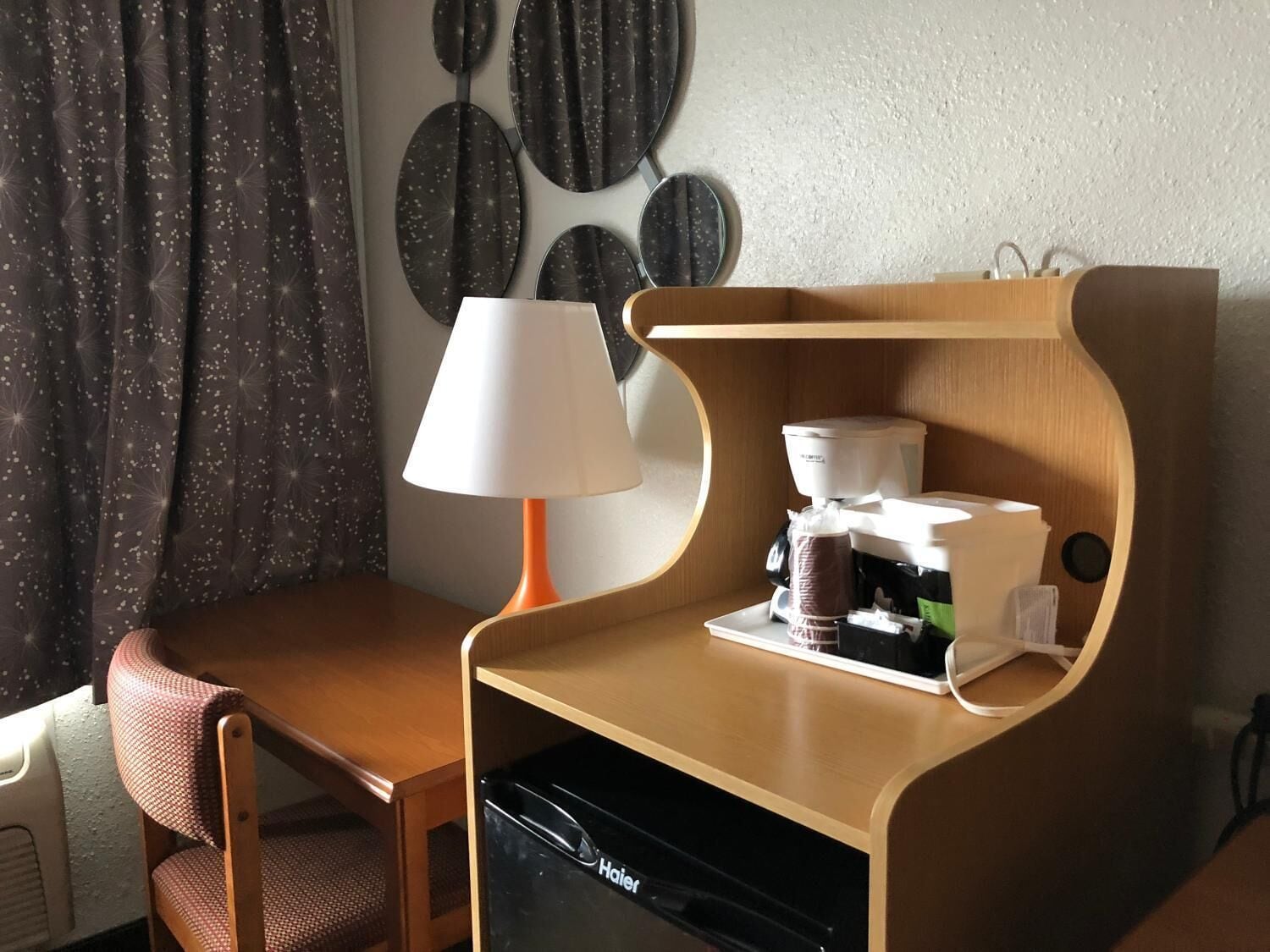A compact hotel room with a wooden desk, a chair, a coffee maker, and a dishwasher.