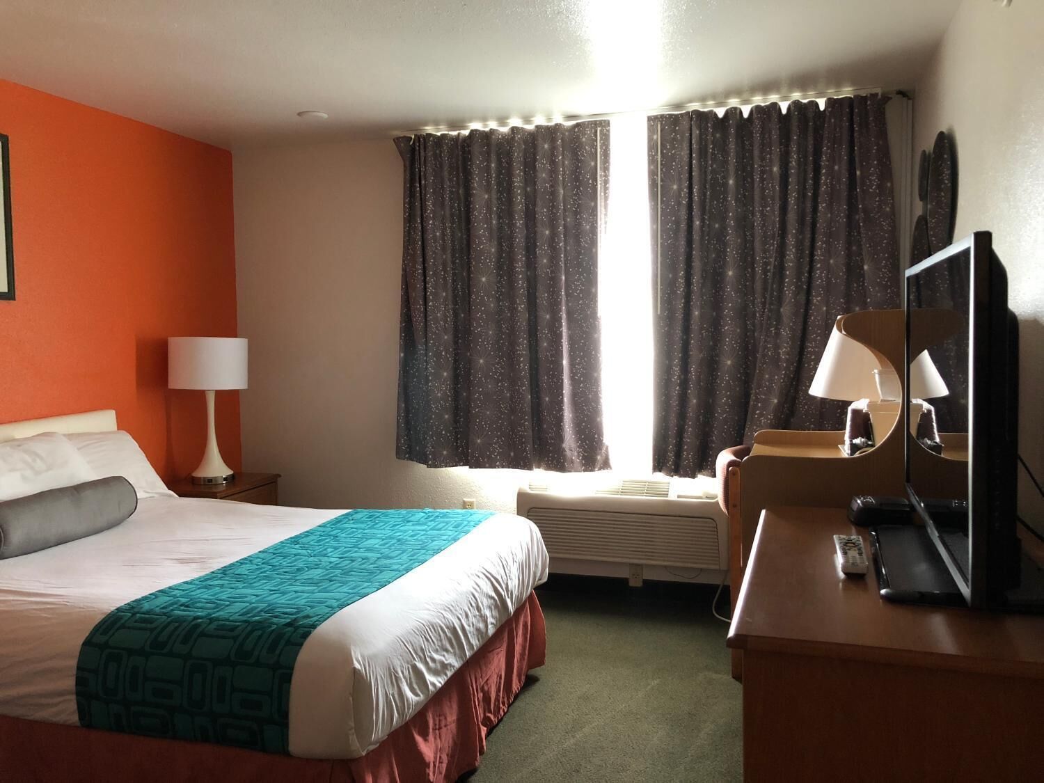 A hotel room with a bed, a desk, a chair, a lamp, a television, and a window with curtains.